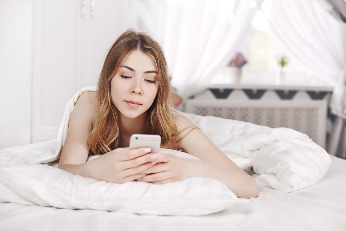 Cute smiling girl looks at the phone, lying on the bed. example image 1