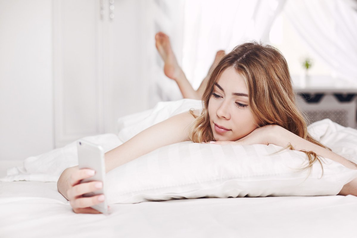 Cute smiling girl looks at the phone, lying on the bed. example image 1
