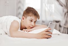 Teen boy with lies on the bed and looks at the phone. Product Image 1