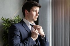 portrait of a businessman in a dark blue suit tying a tie Product Image 1