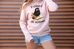 Cute black crow sitting on an open book with soft flowers and sparkles, paired with the phrase “F-CAW-F I’m Reading”