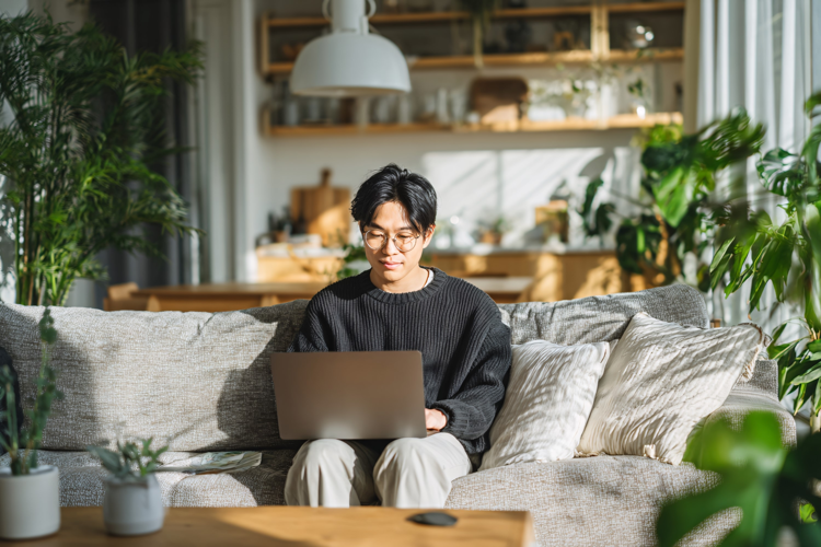 Young Asian Man Working on Laptop on Sofa at Home, Cozy