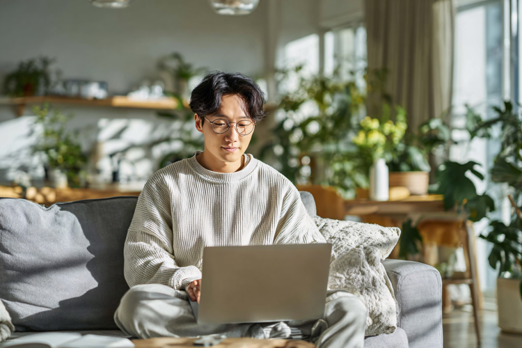Young Asian man working on laptop from home on cozy sofa,